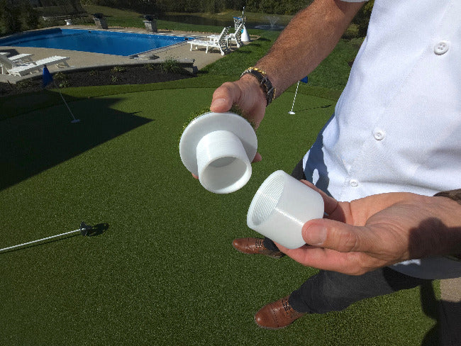 Man holding white and adjustable golf cup in up insert in 2 pieces over top a putting green with pool and pond in the background. 
