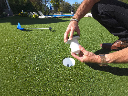 Person adjusting golf cup insert over a golf hole on a putting green with a pool in the background.