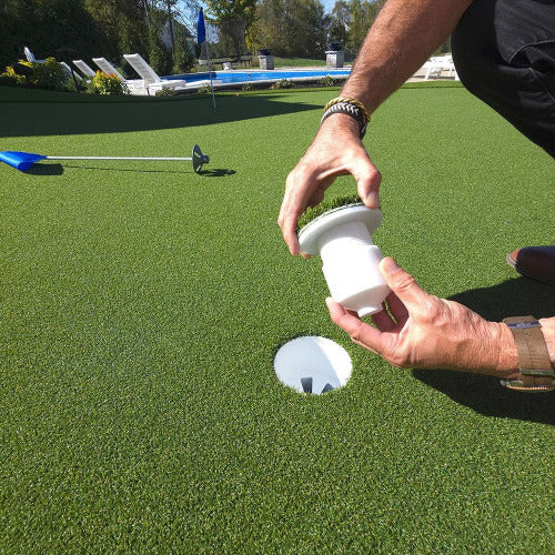 Man holding white Golf cup insert above golf hole with blue flag on putting green a pool in the background